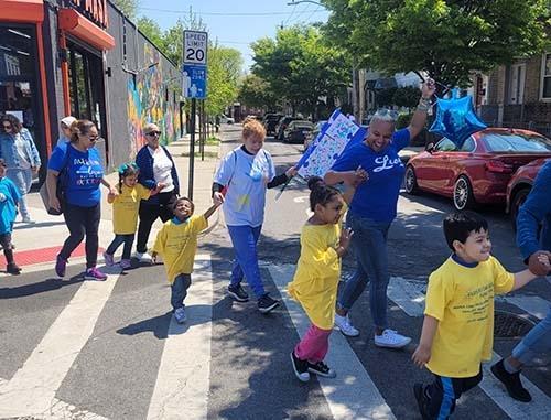 Children in yellow shirts walk across the street as they hold hands with adults in blue shirts.