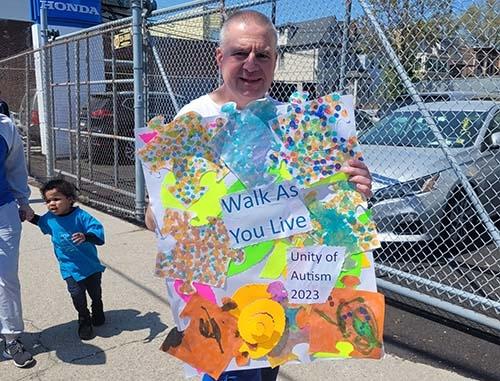 A man holds a poster saying: Walk As You Live. Unity of Autism 2023.