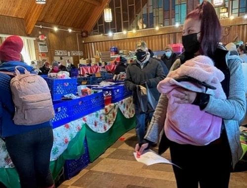 A woman with a mask holds her child. In the background bins of clothes and food are set on a table.