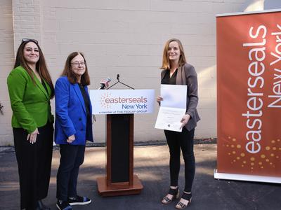 Three women smile toward the camera as they stand in front of a Easterseals New York sign.