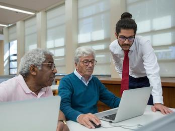 A older man is viewing his computer as two other men lean in to look at his screen.